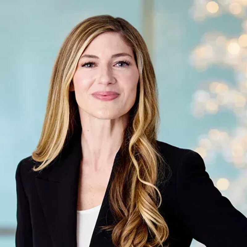 The image shows a woman with long blonde hair, wearing a black blazer over a white top. She has a neutral expression and appears to be in a professional setting. The background is blurred, with a light blue hue and some out-of-focus lights, suggesting an office or studio environment.